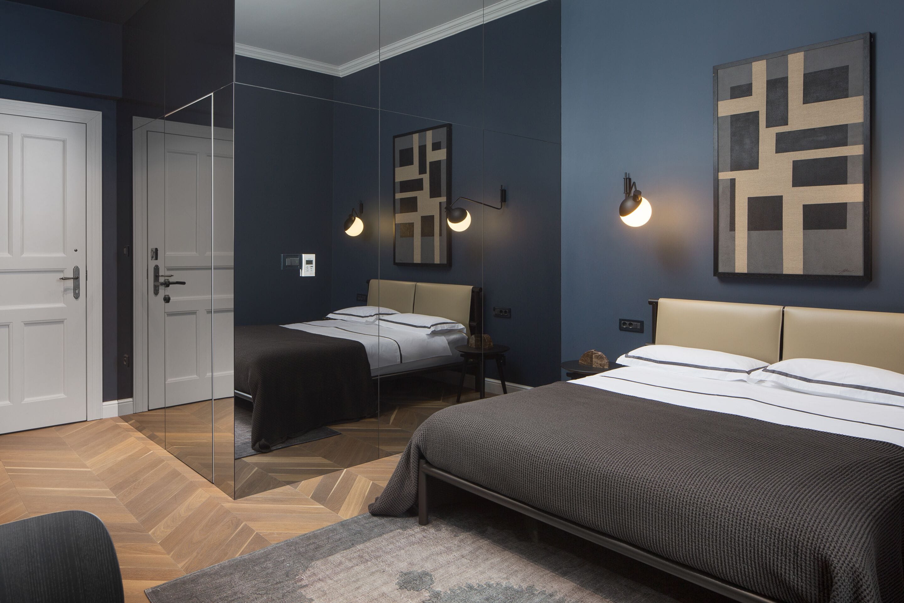 A modern bedroom with a large bed, a smaller bed, and a chair. There are two bedside tables and a mirror.