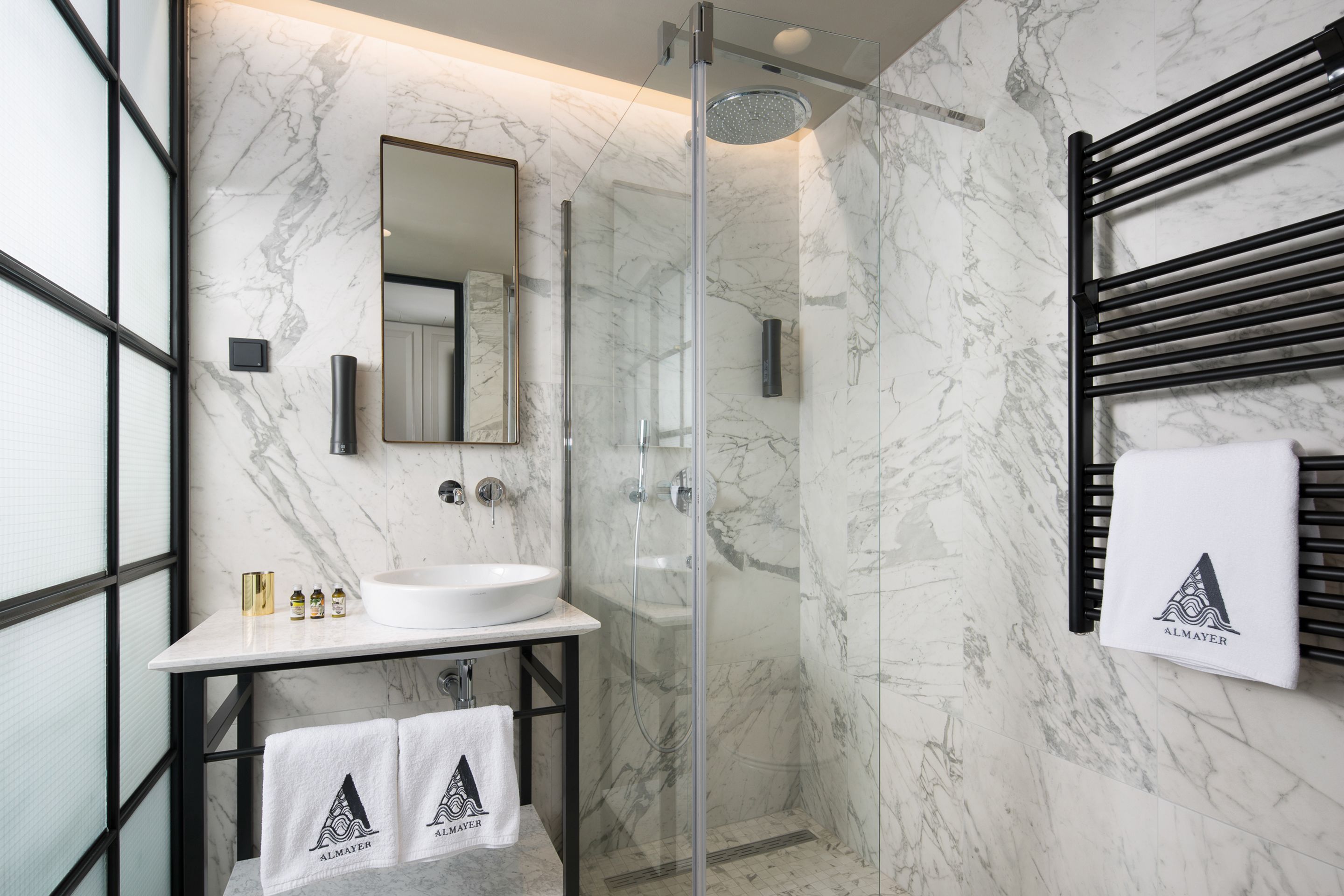 A modern bathroom with a marble wall, a glass-enclosed shower, a white sink, and black towel racks.