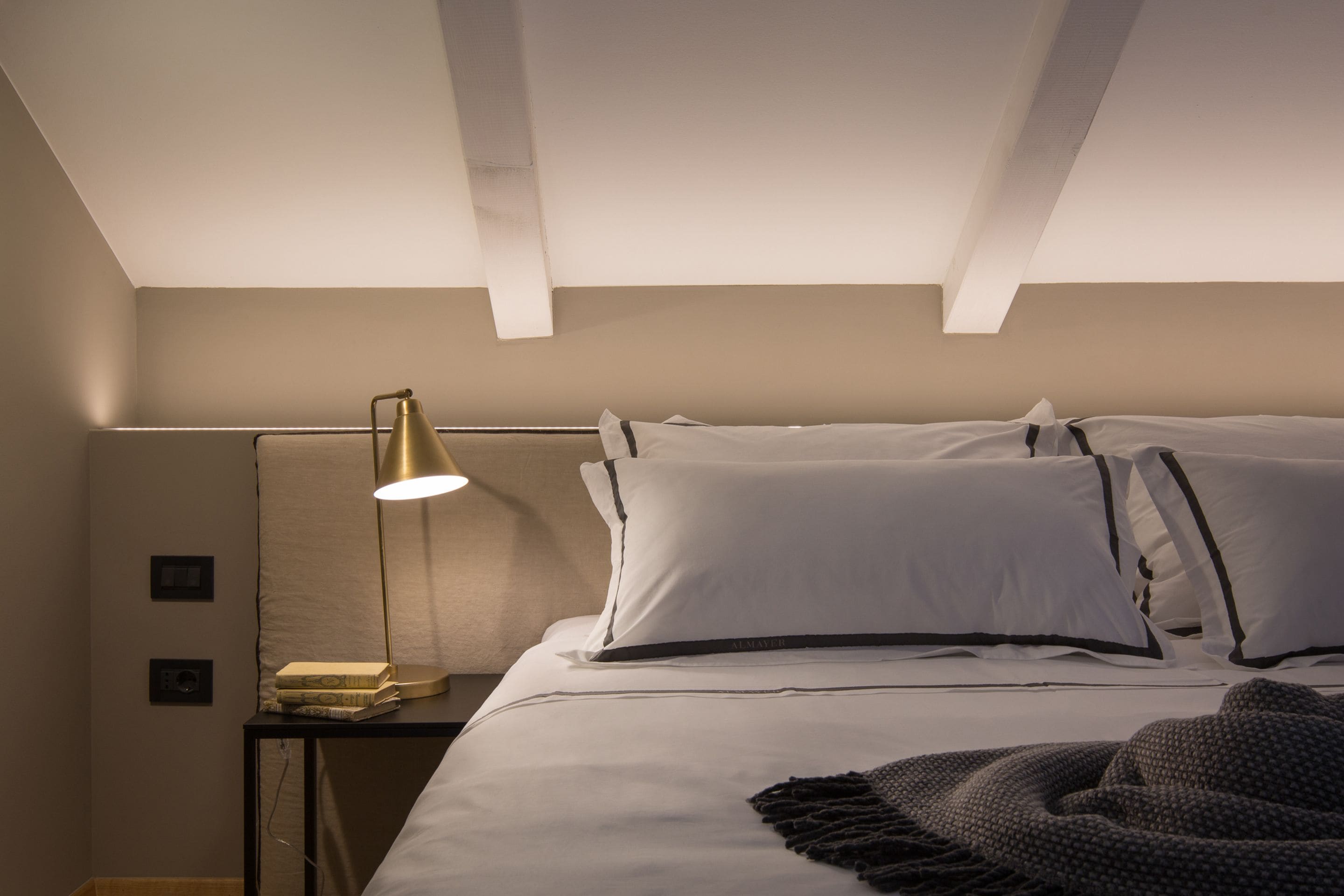 A neatly made bed with white linens and black trim, a bedside lamp, and a small stack of books on a nightstand.