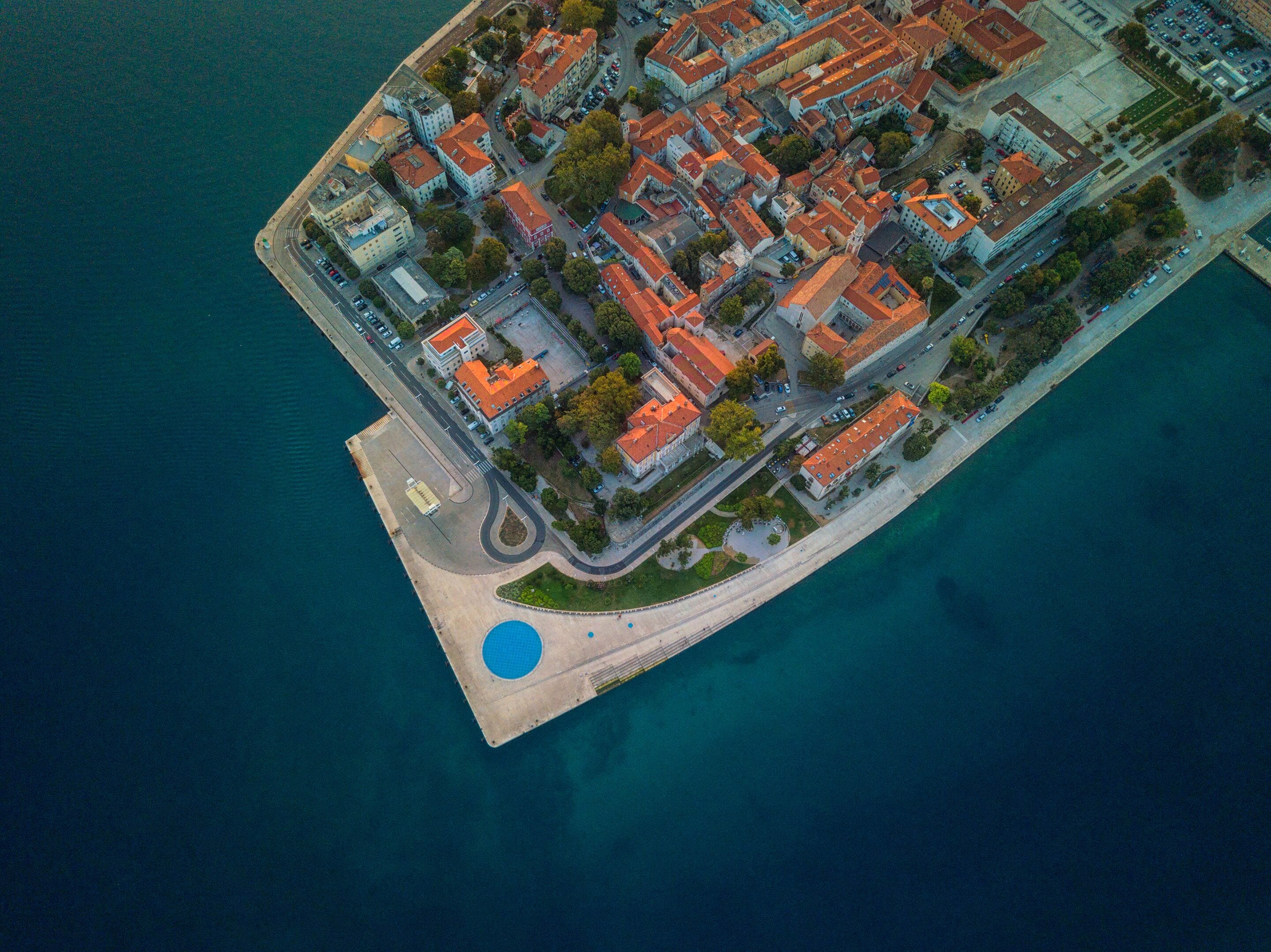 An aerial view of a coastal town with a mix of residential and commercial buildings, a prominent waterfront promenade, and a large blue public pool.