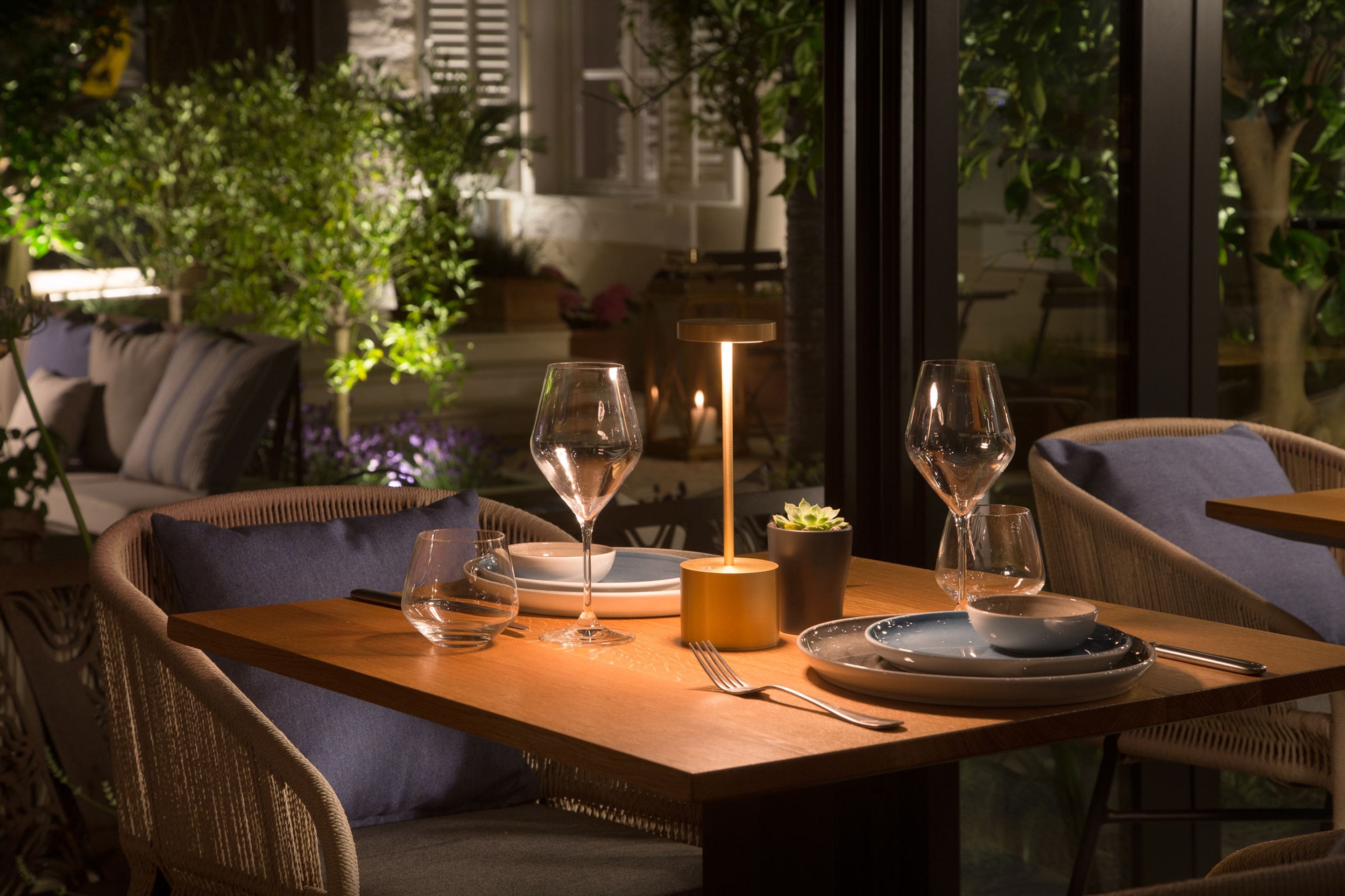 A dining table set with plates, glasses, and a candle, surrounded by wicker chairs in a well-lit indoor space with plants and a view through large windows.