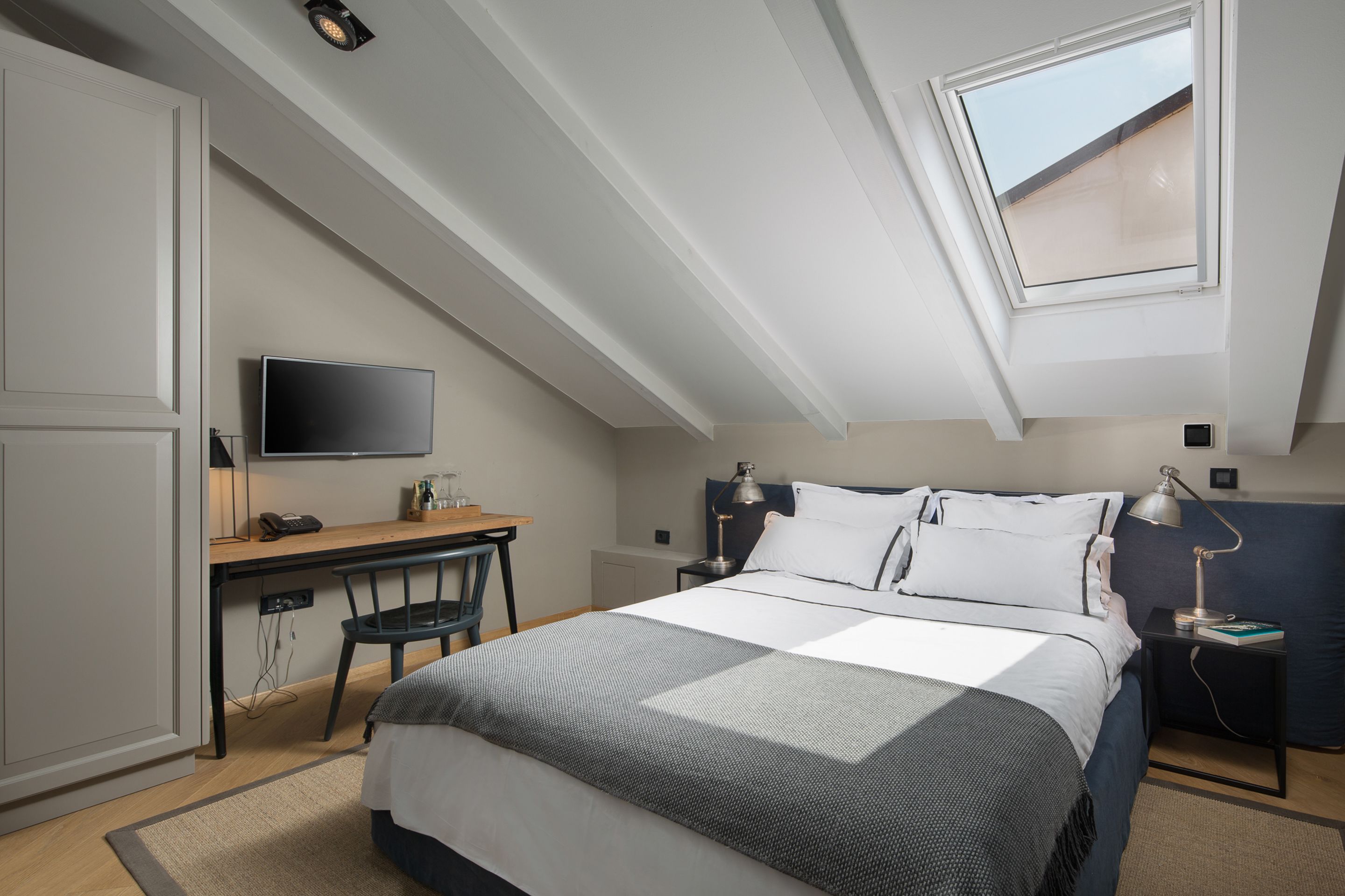 A bedroom with a bed, desk, chair, TV, and a skylight.