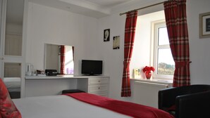 Quarto de hotel com cama, escrivaninha com computador, cadeira, janela com cortinas e radiador.