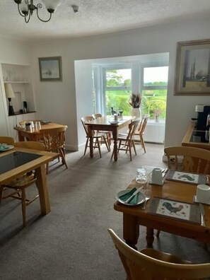 A dining area with wooden tables and chairs, a chandelier, and a view through a large window. - Airds Farm Guest House (Castle Douglas)