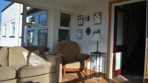 Sala de estar com sofá, poltrona e abajur. Há quadros nas paredes e uma janela com persianas.