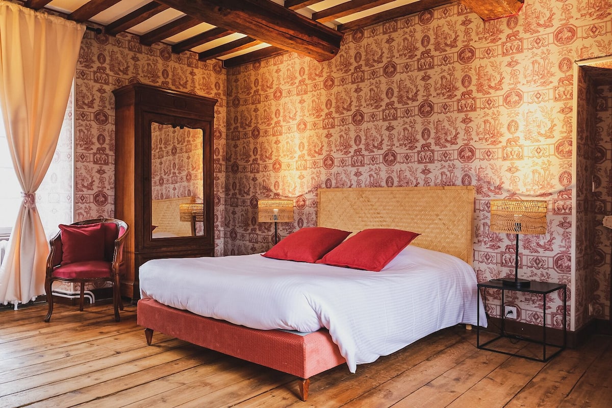 A bedroom with a large bed, two red pillows, a wooden floor, a chair, and patterned wallpaper.