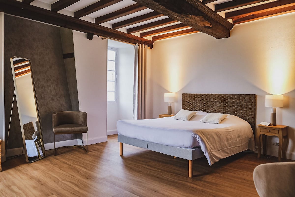 A bedroom with a bed, bedside lamps, a chair, a mirror, and wooden beams on the ceiling.