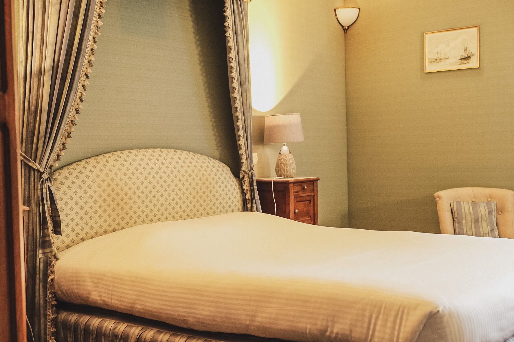 A neatly made bed with a headboard, a bedside table with a lamp, and a chair in a room with a simple wall and a framed picture.