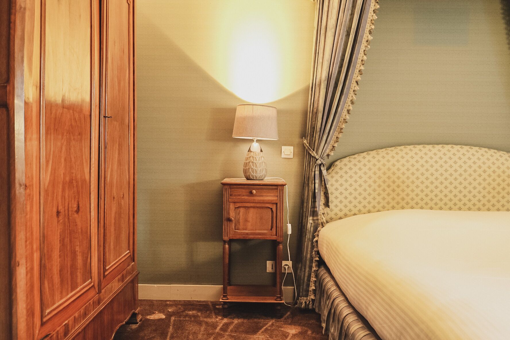 A bedroom with a wooden wardrobe, a bedside table with a lamp, and a bed with a patterned bedspread.