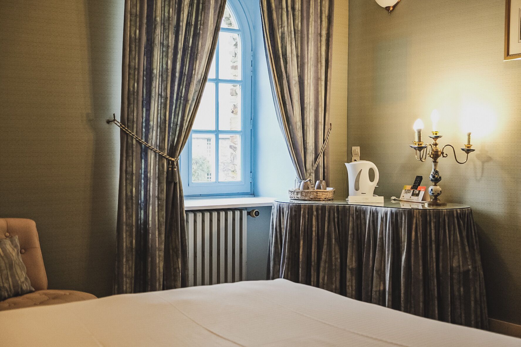 A hotel room with a large bed, a table with a kettle and a tray, curtains, and a window with a view.