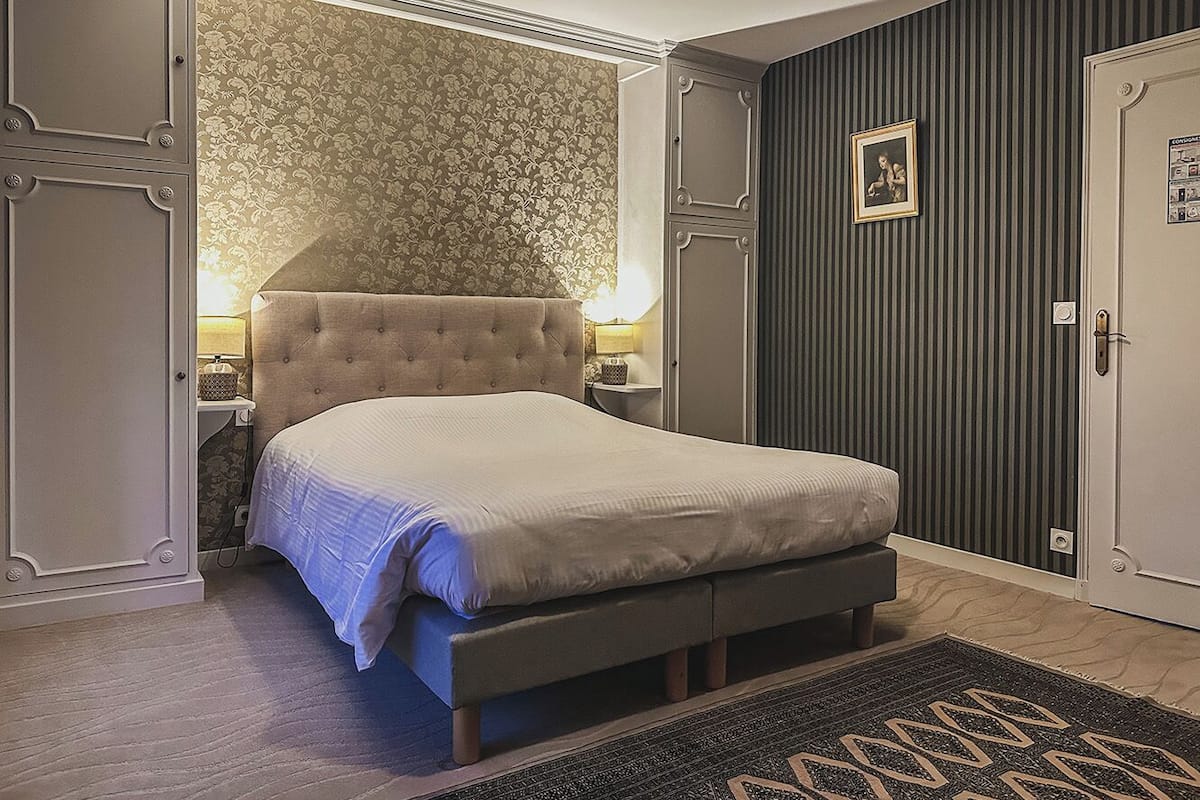 A bedroom with a tufted headboard, a bed with a white comforter, a bedside table with a lamp, a dark striped wall, and a door with a picture.