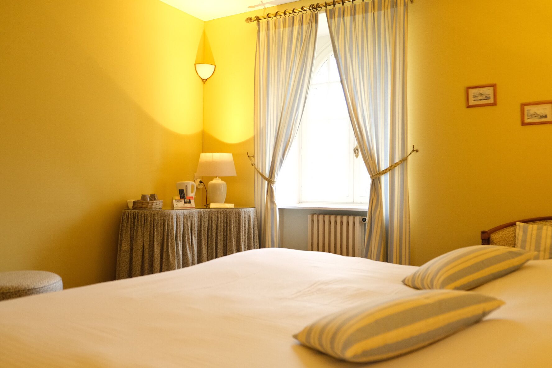 A hotel room with a bed, a bedside table with a lamp, a window with curtains, and a radiator.