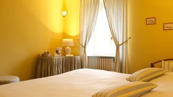 A hotel room with a bed, a bedside table with a lamp, a window with curtains, and a radiator.