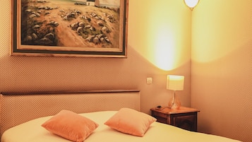 Una habitación de hotel con cama, dos almohadas, un cuadro de una escena costera, una mesita de noche con lámpara y una pared con textura.