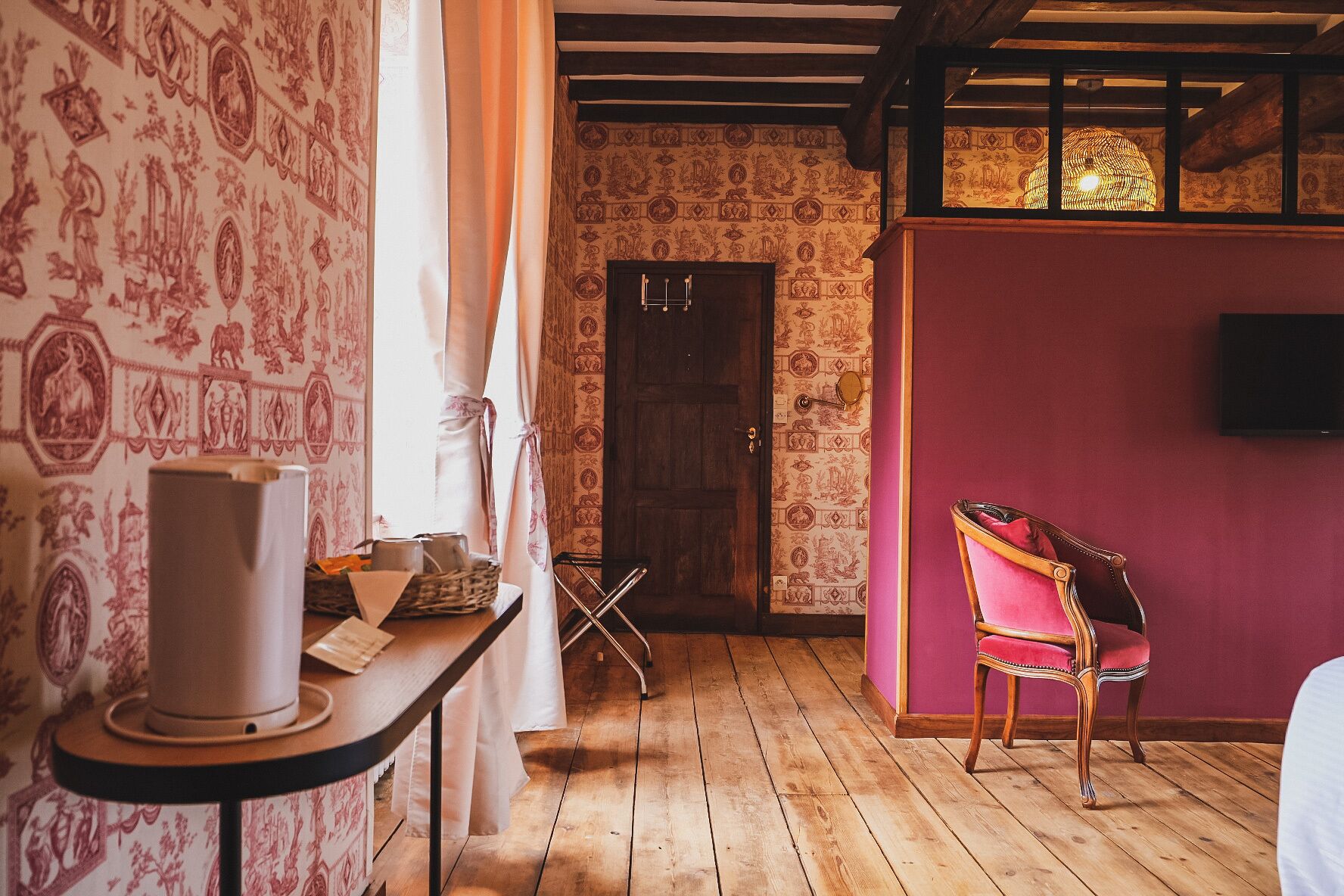 Een kamer met behang met patroon, een houten vloer, een rode stoel, een tafel met een lamp en een deur.