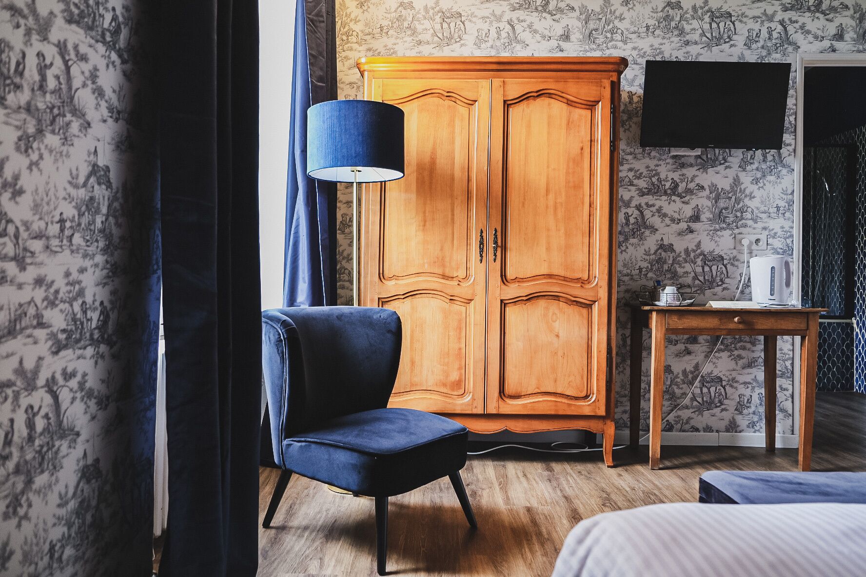 A room with a wooden wardrobe, a blue chair, a small table with a kettle, and a bed.