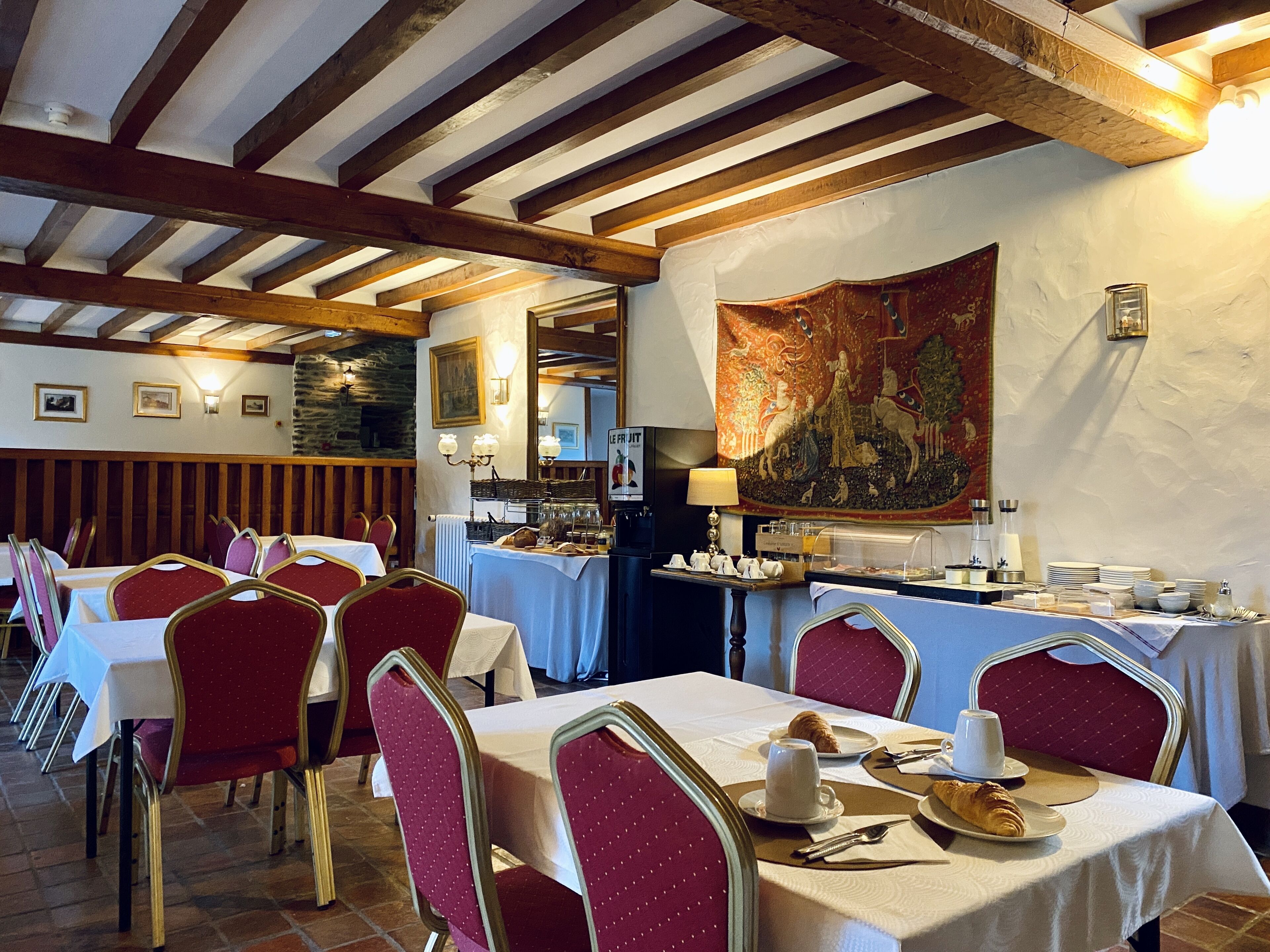 A restaurant with wooden beams, tables with white tablecloths, red upholstered chairs, and a tapestry on the wall.