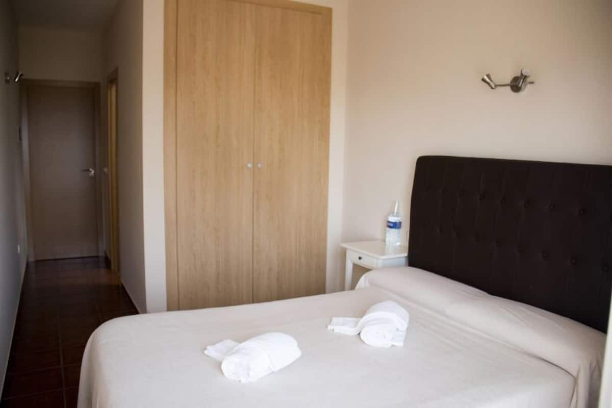 Quarto de hotel com cama, guarda-roupa de madeira, mesa de cabeceira com uma garrafa e luminária na parede.