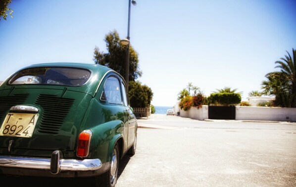 A vintage green car parked on a sunny street with a clear view of the ocean in the background.