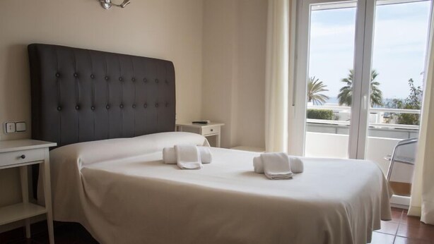 Hotelkamer met een groot bed, een nachtkastje, een kleine tafel en uitzicht op palmbomen en een stadsgezicht door het raam.
