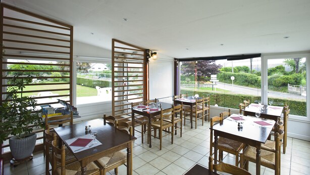 A restaurant with multiple tables and chairs, large windows with a view of a garden and street.