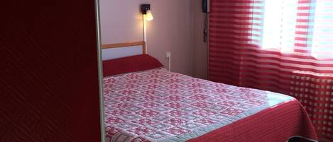 A bedroom with a bed, a wall-mounted lamp, a telephone, and a window with red curtains.