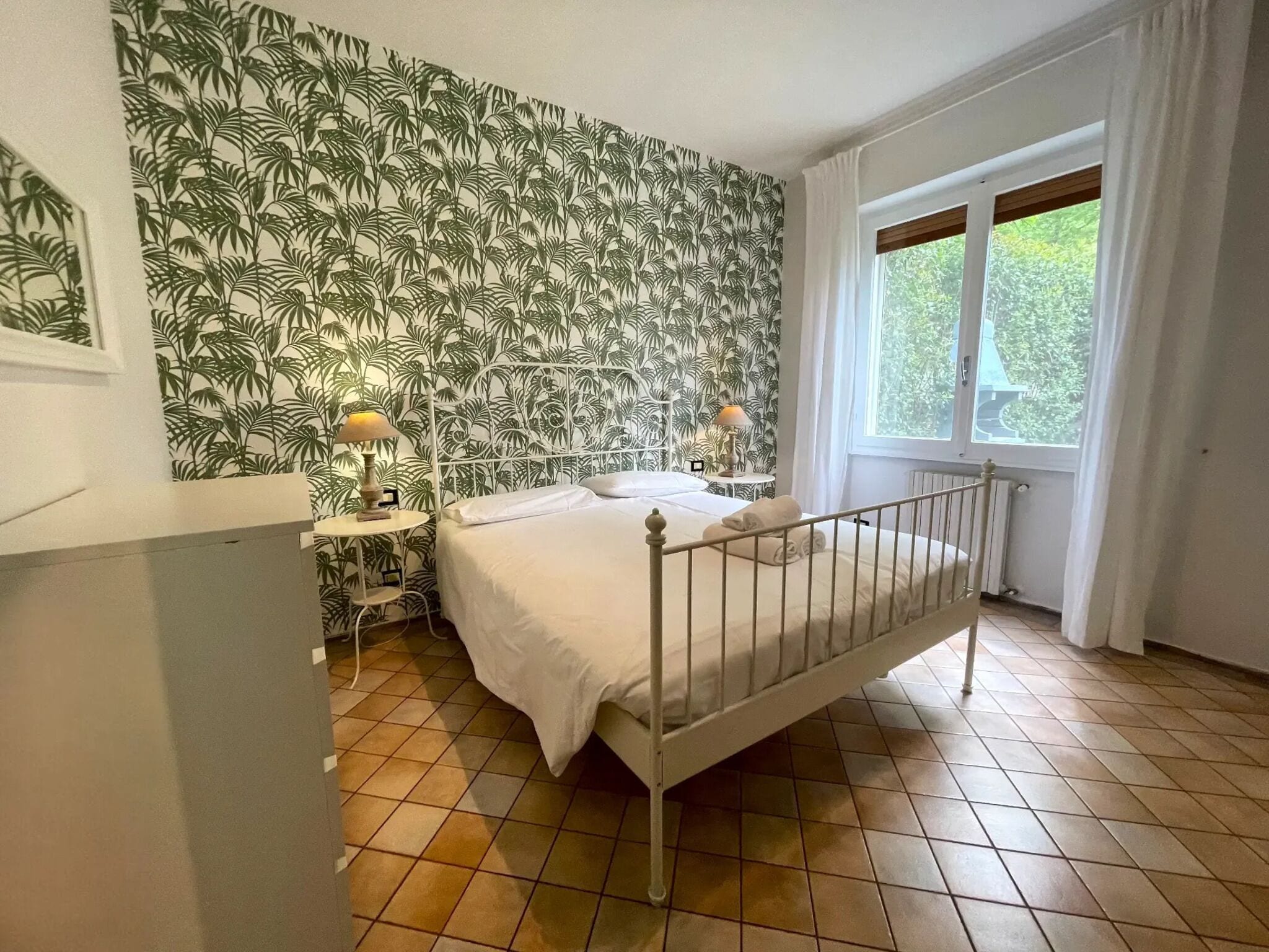 A bedroom with a bed, two bedside tables, a window with curtains, and a wall with a tropical leaf pattern.