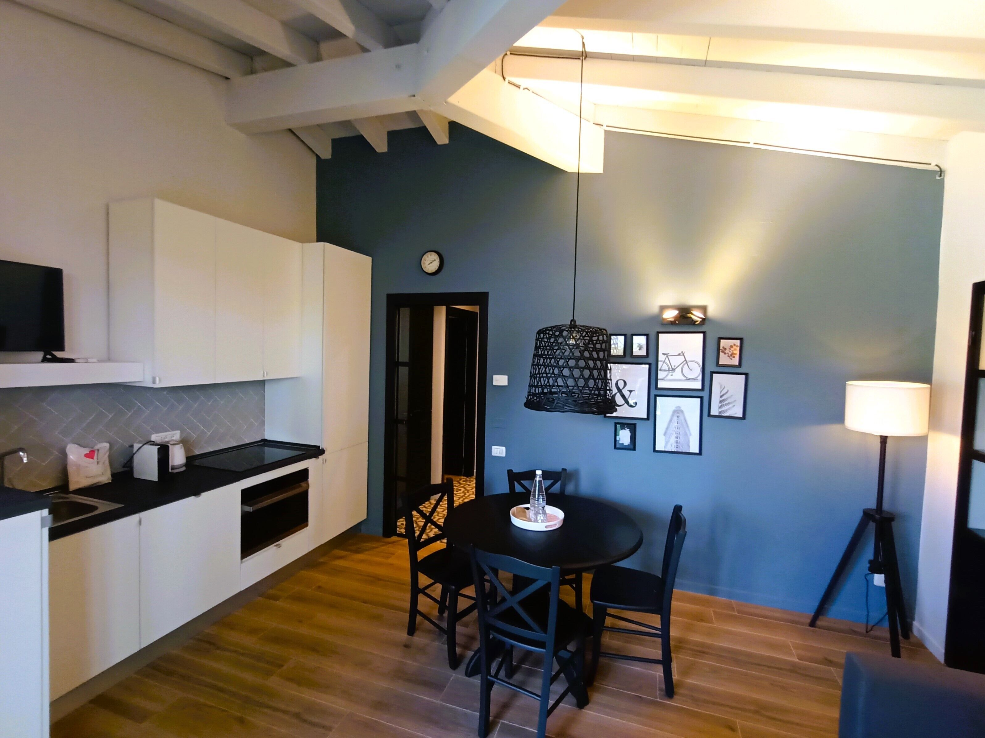 A modern kitchen with white cabinets, a black countertop, and a dining area with a round table and chairs. There is a hanging light fixture above the table and a clock on the wall.