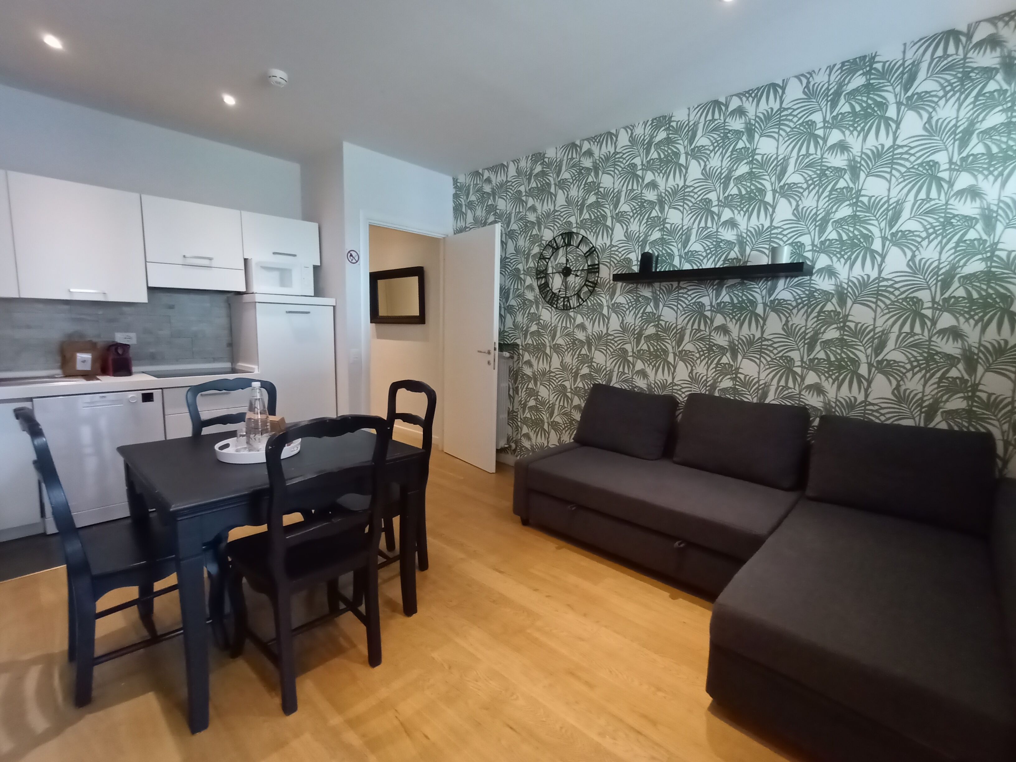 Un ambiente compatto con angolo cottura, zona pranzo con tavolo e sedie, e divano addossato a una parete decorata con motivo tropicale.