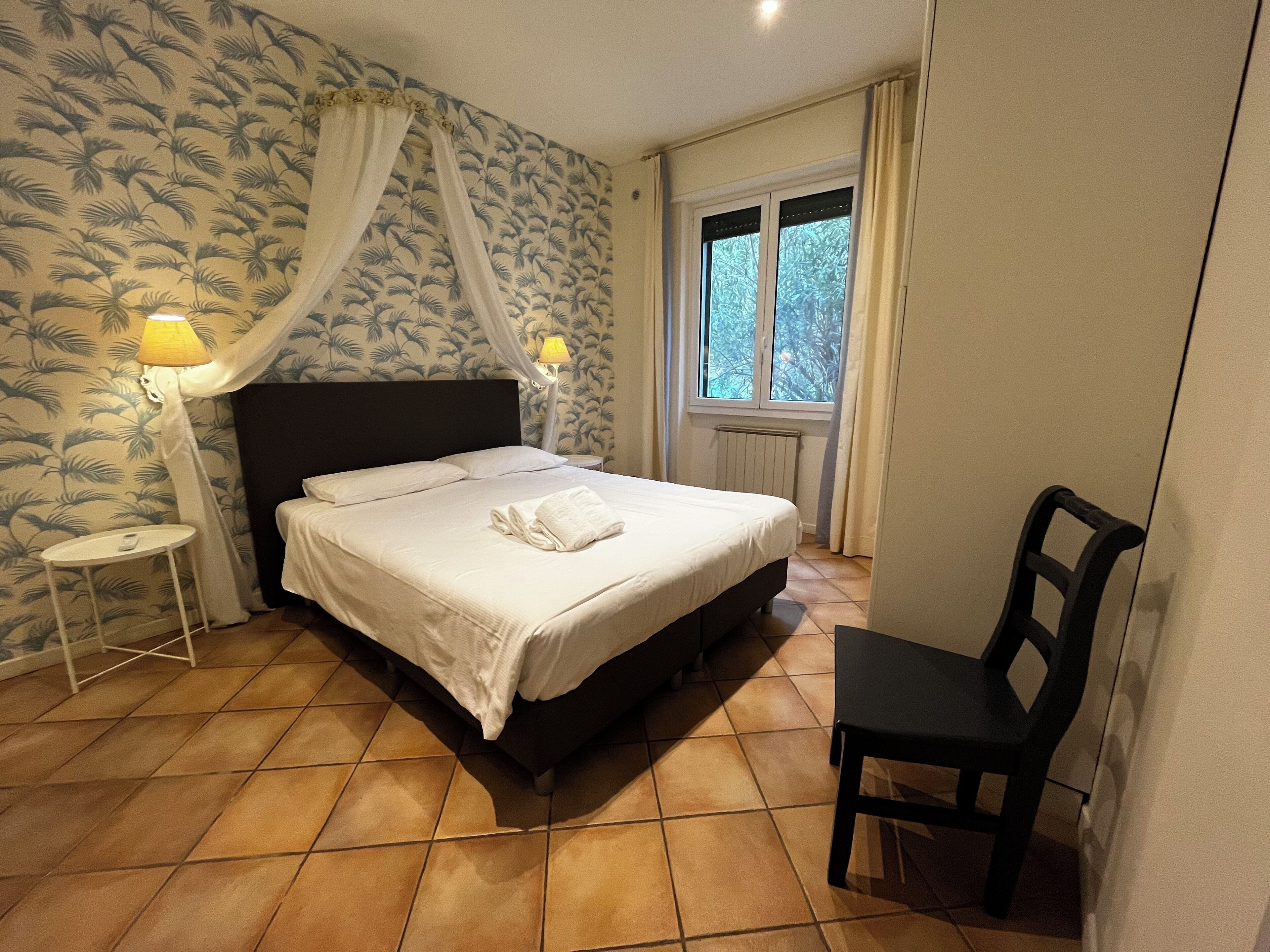 A bedroom with a bed, a chair, a nightstand, a lamp, and a window with curtains.