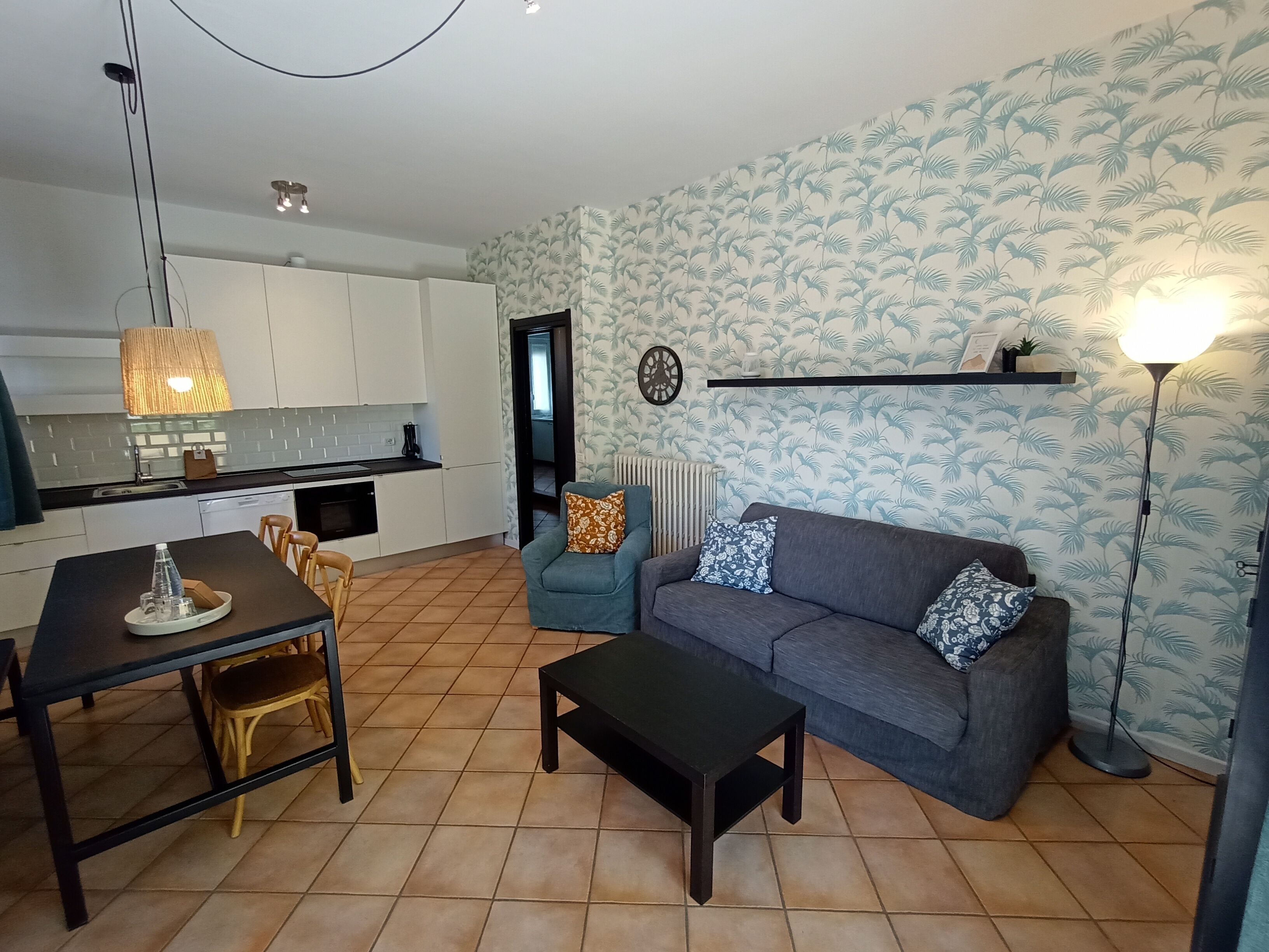 Een moderne woonkamer met een grijze bank, een eettafel met stoelen, een zwarte salontafel en een keuken met witte kasten en een hangende lamp.