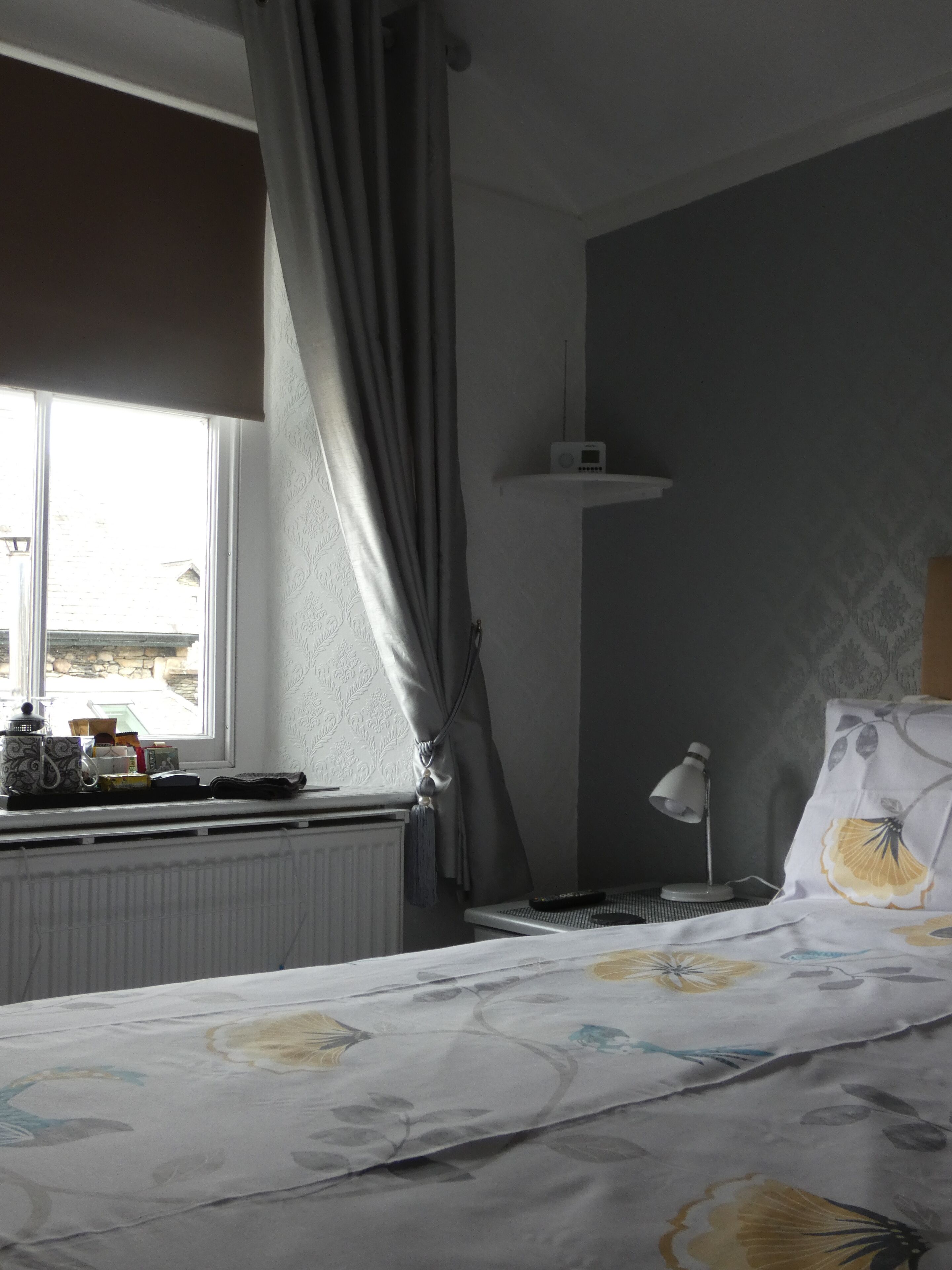 Une chambre d’hôtel avec un lit, une fenêtre avec des rideaux, un radiateur, une table de chevet avec une lampe et une radio.