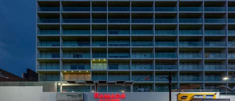 Un hotel de varios pisos con un llamativo letrero 'Ramada', que presenta un diseño arquitectónico moderno con balcones y techo plano.