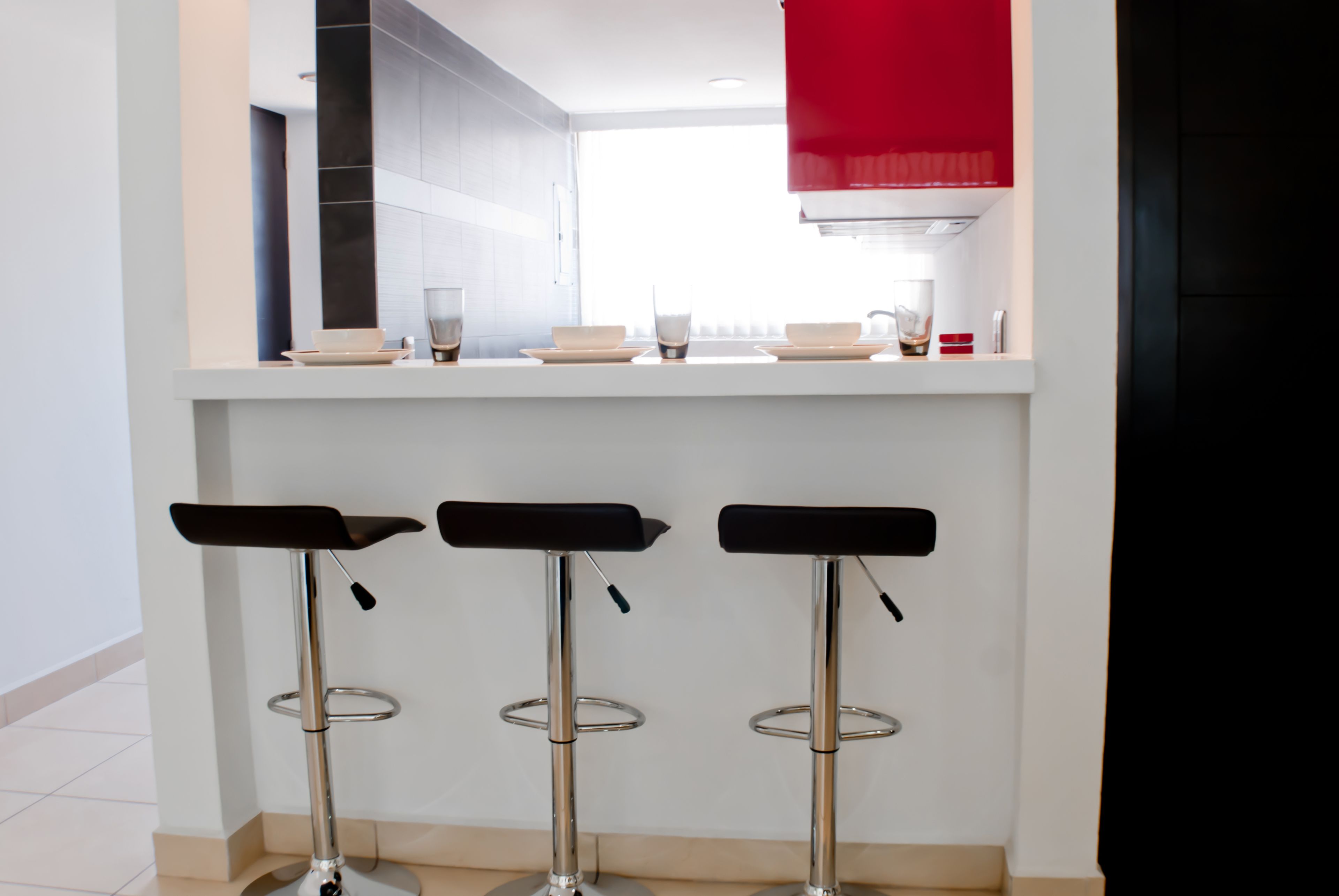 A modern kitchen with three bar stools, a red hanging light, and a countertop with dishes.