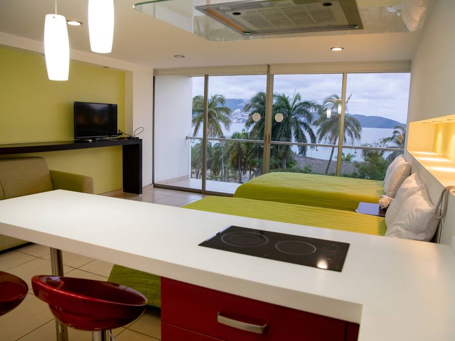 A modern hotel room with a kitchen area, a bed with a green bedspread, and a balcony with a view of palm trees and mountains.