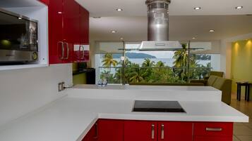 Una cocina moderna con armarios rojos, un microondas de acero inoxidable y un amplio ventanal con vistas a un paisaje tropical.