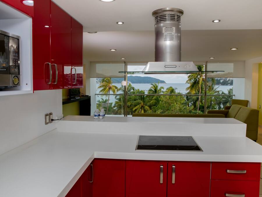 A modern kitchen with red cabinets, a stainless steel microwave, and a large window overlooking a tropical landscape.