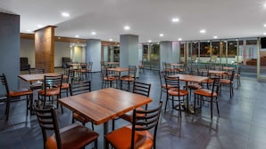 A spacious indoor dining area with multiple wooden tables and chairs, large windows offering views of the outdoors, and a modern architectural design with exposed beams and a high ceiling.