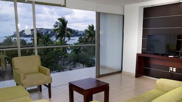 A modern living room with a large window offering a view of a coastal area, a flat-screen TV, and a wooden entertainment unit.