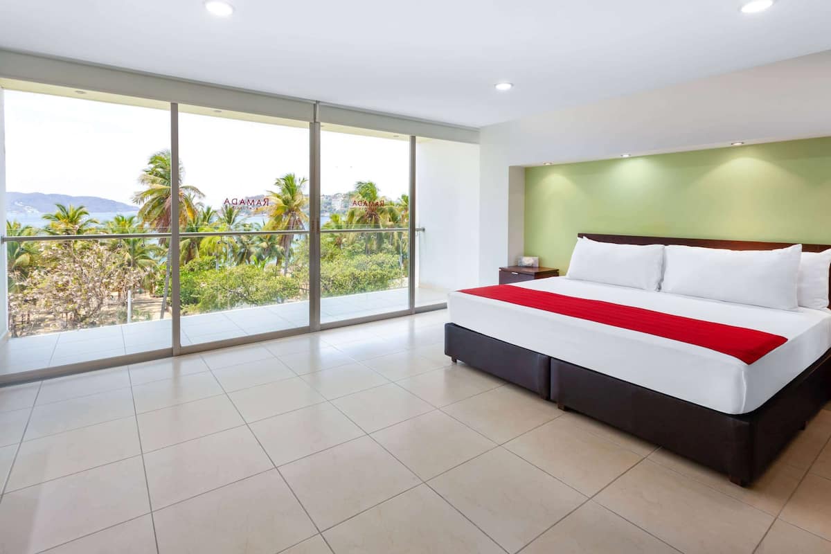 Een moderne hotelkamer met een groot bed, een nachtkastje en uitzicht op palmbomen en bergen door middel van kamerhoge ramen.