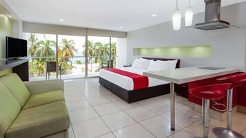 Quarto de hotel moderno com cama, escrivaninha, televisão e varanda com vista para palmeiras e o mar.