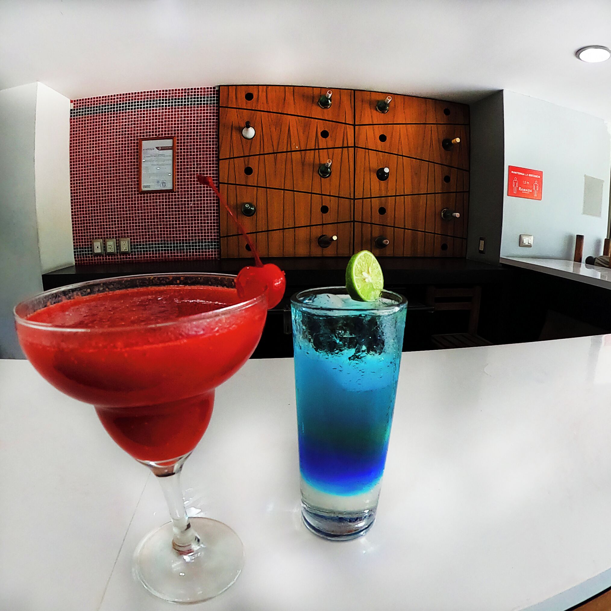Zona de bar con un cóctel rojo y una bebida azul, pared de azulejos y panel de madera con un letrero.
