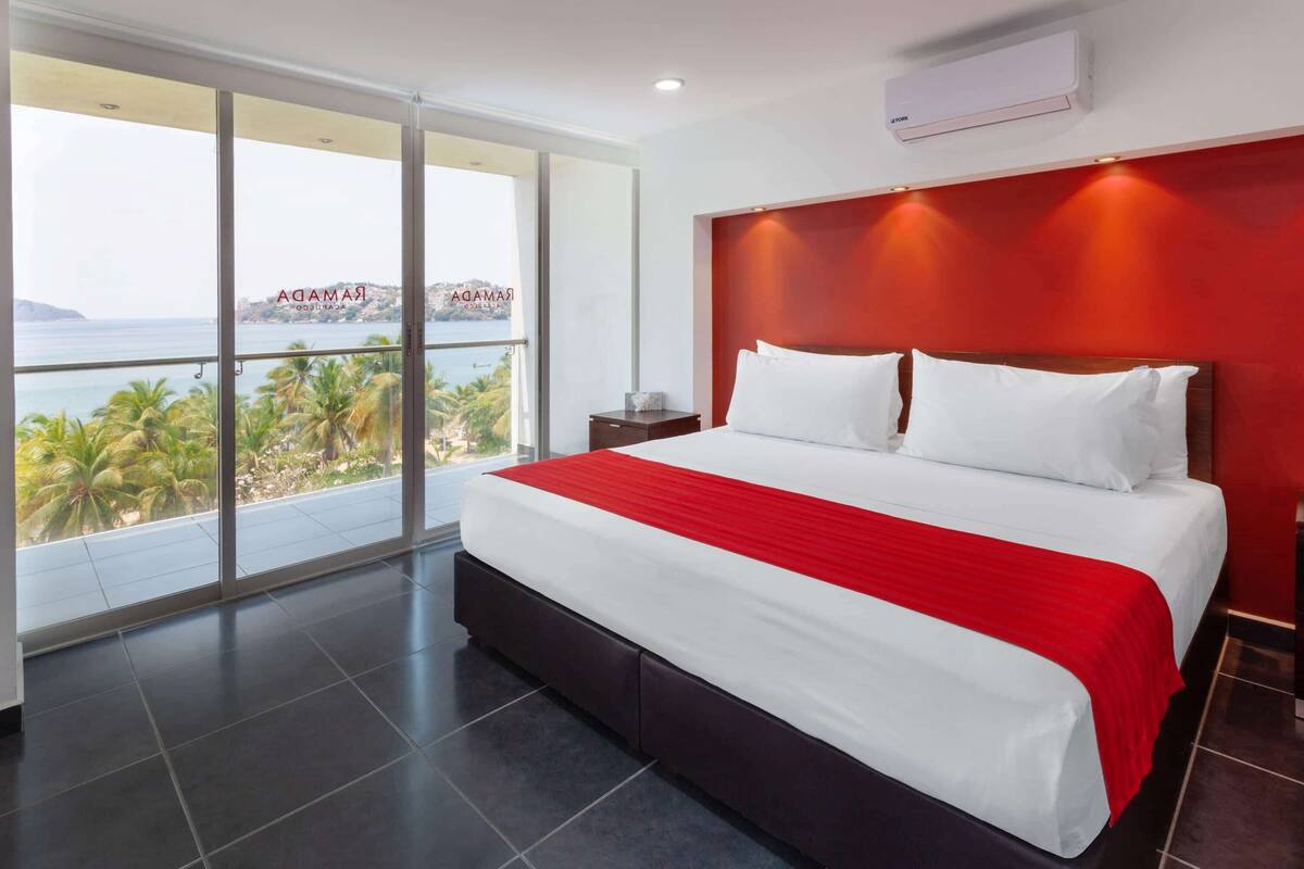Een hotelkamer met een groot bed, een rode hoofdbord en uitzicht op palmbomen en de zee.