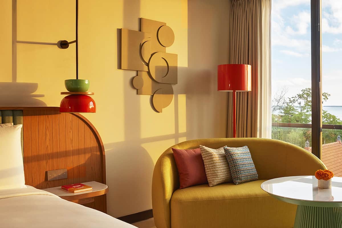 A hotel room with a bed, a sofa, a small table with a vase, a wall art piece, and a view of the outdoors.
