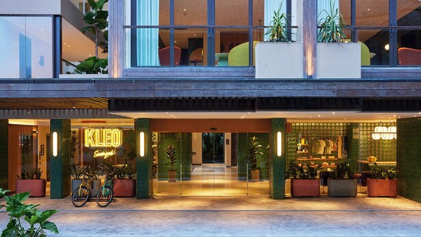 Entrada de um hotel moderno com letreiro de neon, portas de vidro e uma bicicleta estacionada.