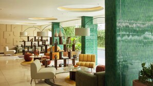 Um lobby moderno com área de estar central, estante de livros e uma grande janela com vista para o verde.
