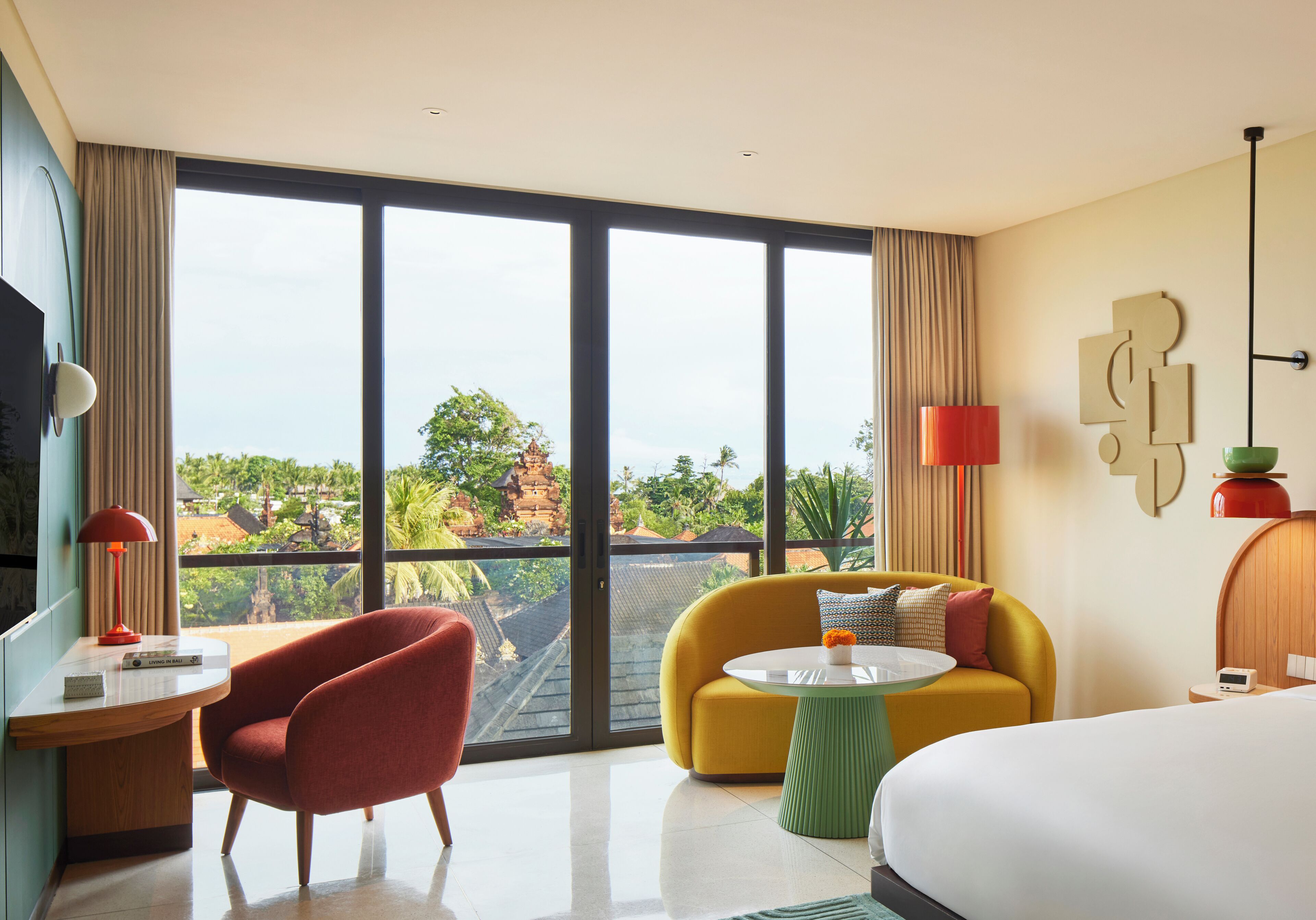 A modern hotel room with a large window, a sofa, a bed, and a desk with a lamp.