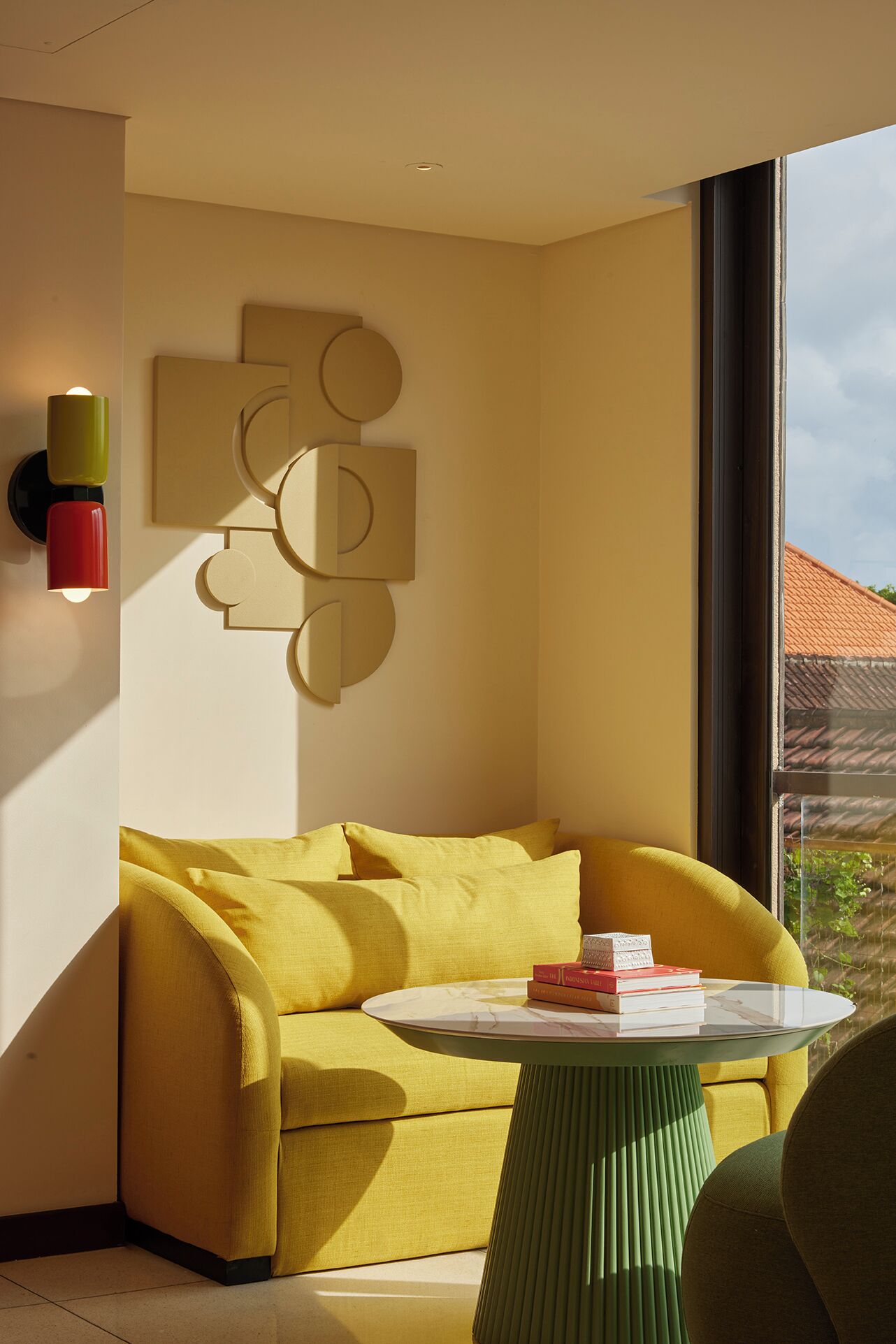 A modern living room with a yellow sofa, a round table with books, and a geometric wall art piece.