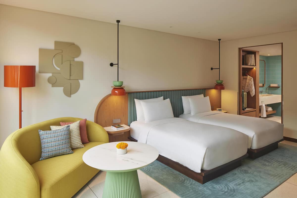 A hotel room with a bed, a sofa, a small table with a vase, a lamp, and a wall-mounted artwork.