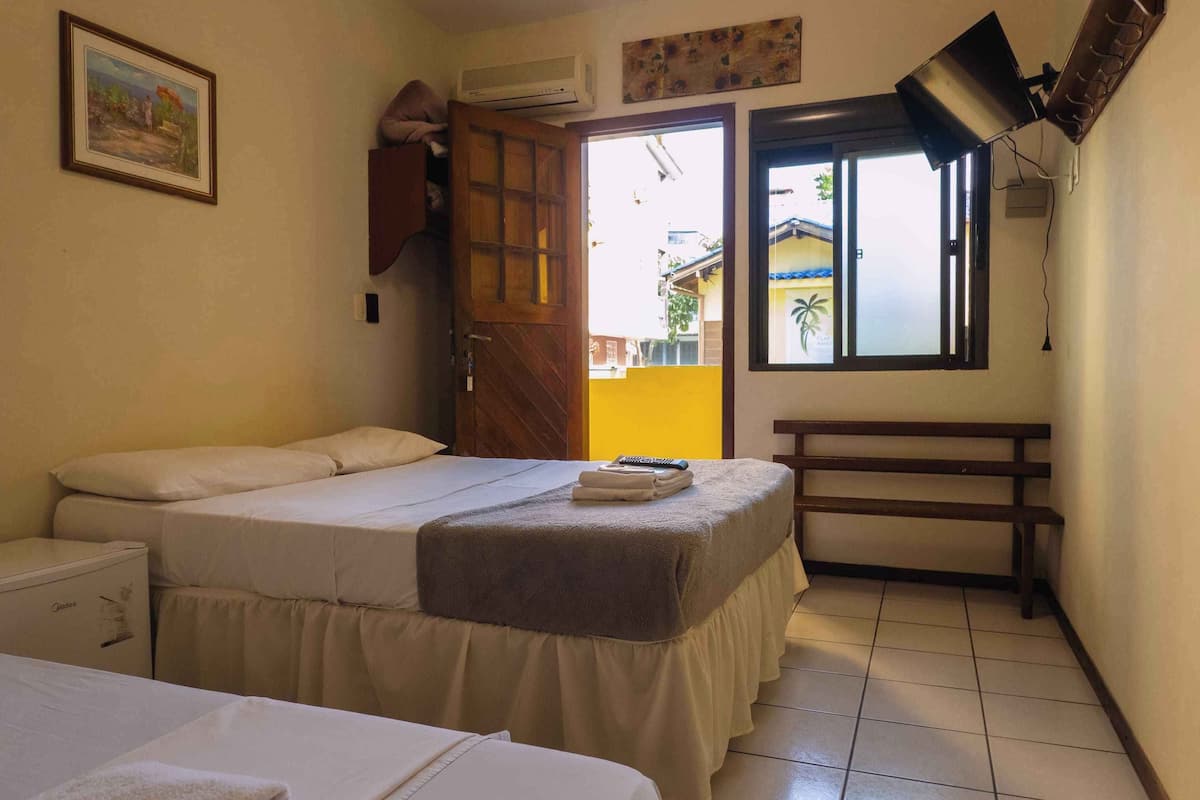 A hotel room with two beds, a bench, a door leading outside, and a window with a view of a building.